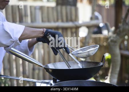 Ein professioneller Koch mit schwarzen Handschuhen verwendet eine Zange und ein Drahtsieb, um Speisen in einem heißen Wok zu braten – während einer authentischen, traditionellen Outdoor-Küche Stockfoto