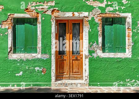 Ein altes Haus in Burano, Italien, mit leuchtend grüner abblätternder Farbe, die rote Ziegelsteine zeigt, einer klassischen Holztür und Fensterläden Stockfoto