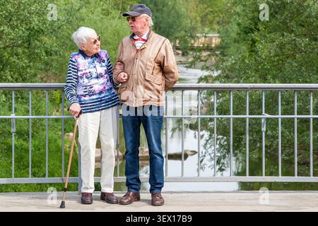 Ein älteres Paar, das Arm in Arm über eine Brücke geht, die ältere Frau mit einem Stock, während sie mit ihrem Partner redet. Stockfoto