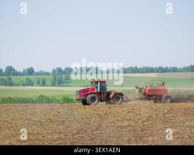 Roter Traktor zieht landwirtschaftliche Maschinen über fruchtbares Feld zur Bodenbearbeitung. Bauer arbeitet auf dem Hof während der Pflanzsaison. Industrielles Land Stockfoto