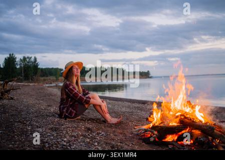 Eine Frau mit Strohhut und kariertem Hemd nimmt ruhige Momente an einem Kiesstrand wahr. Sie sitzen barfuß am warmen Lagerfeuer und reflektieren sich, während Sie den wunderschönen Sonnenuntergang über dem ruhigen Seewasser beobachten Stockfoto