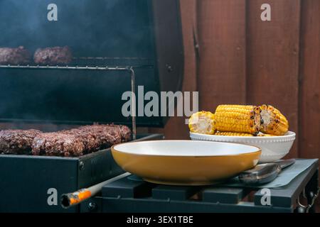 Gartengrill mit gegrillten Burgern und Maiskolben auf Holzkohlegrill, fertig zum Essen. Die Outdoor-Szene fängt ein entspanntes Sommerkochen ein Stockfoto