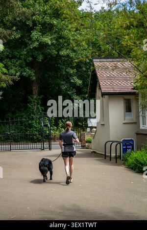 Person, die mit einem schwarzen Hund auf einem gepflasterten Weg neben einem kleinen Gebäude mit einem gekachelten Dach joggt. Die Outdoor-Szene fängt einen aktiven Lebensstil ein Stockfoto