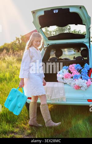 Junge Frau mit einem stilvollen türkisfarbenen Picknickkoffer neben einem blumengefüllten Kofferraum während der goldenen Stunde auf dem Land, die einen Sommerurlaub genießt Stockfoto