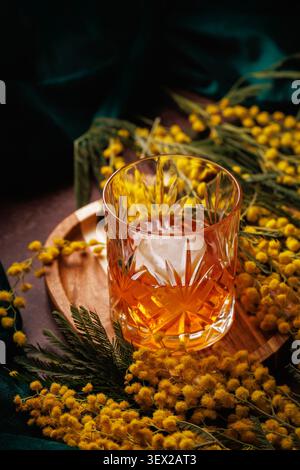 Kristall-Whiskyglas mit Eis und Mimosenblüten auf Holztablett Frühlingsgetränk Stockfoto