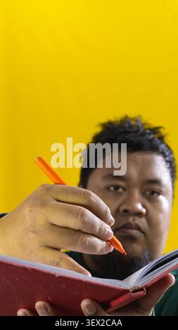 Porträt eines asiatischen Mannes in grünem Hemd, der in rotem Buch schreibt, Fokus auf Stift mit verschwommenem Gesicht, gelber Hintergrund, der Kreativität und Studium repräsentiert. Stockfoto