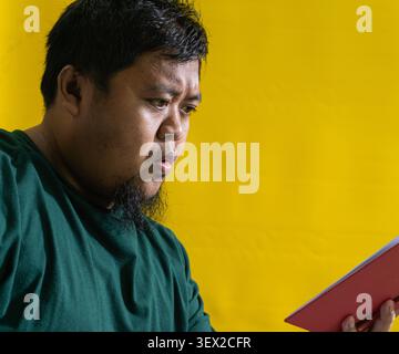 Asiatischer Mann im grünen Hemd liest rotes Buch, überraschte Reaktion vor gelbem Hintergrund, Entdeckungskonzept und Lernneugier. Stockfoto