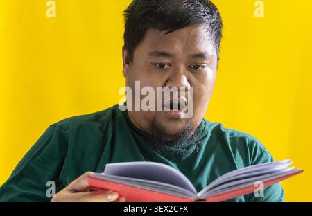 Asiatischer Mann im grünen Hemd liest rotes Buch, überraschte Reaktion vor gelbem Hintergrund, Entdeckungskonzept und Lernneugier. Stockfoto