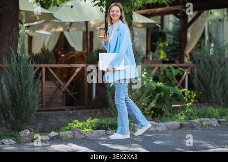 Junge Frau, die draußen mit Kaffee und Laptop läuft Stockfoto
