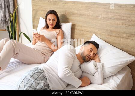 Unzufriedene junge Frau mit Handy und ihr schlafender Ehemann im Schlafzimmer Stockfoto