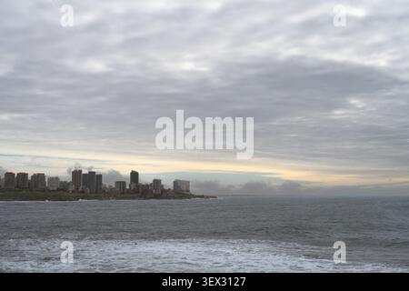 Graue Wolken erheben sich über der fernen Skyline der Stadt. Schneebedecktes Wasser reicht bis zum Horizont. Gebäude reflektieren das sanfte Licht. Ruhige, winterliche Szene. Ideal für Stockfoto