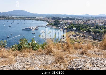 Panoramablick auf die Küste von korfu, griechenland, mit einer historischen Festung, charmantem Hafen mit Booten und fernen Bergen. Stockfoto