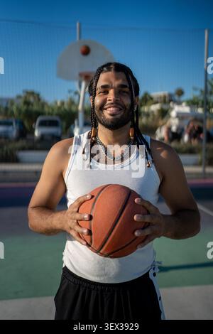Selbstbewusster junger Mann mit Zöpfen, der Basketball auf dem Platz im Freien hält Stockfoto
