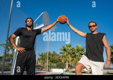 Junge männliche Freunde posieren mit Basketball auf dem Platz Stockfoto