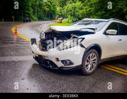 Ein weißer SUV wurde bei einem Autounfall an einem regnerischen Tag auf einer rutschigen Asphaltkurve schwer beschädigt, umgeben von grünen Waldbäumen. Autoversicherung und Traffi Stockfoto