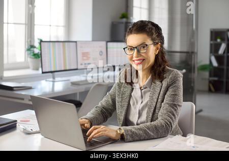 Lächelnde Buchhalterin, die an einem Laptop arbeitet, um Finanzdaten in einem hellen Büro zu analysieren Stockfoto