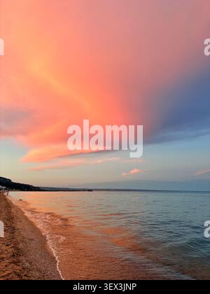 Atemberaubende Küstenlandschaft mit dramatischen rosa und orangen Sonnenuntergangswolken, die sich auf dem ruhigen Meerwasser und der Sandküste spiegeln. Stockfoto