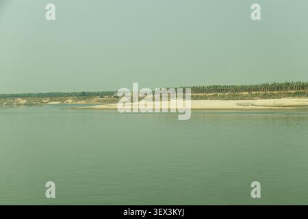 Eine ruhige Flusslandschaft mit einem ruhigen Fluss, einem breiten Sandufer und einer Ansammlung ländlicher Hütten, die sich ruhig zwischen dichten grünen Bäumen unter einem weichen, Stockfoto