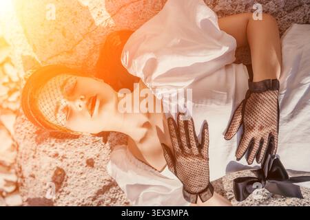 Die Vintage-Sonnenstrahlen der Frau ruhen friedlich auf robustem Stein in einem eleganten weißen Kleid und Spitzenhandschuhen zur goldenen Stunde Stockfoto
