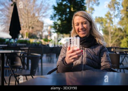 Frau lächelt, sitzt in einem Café im Freien und verbindet sich mit ihrem Smartphone Stockfoto