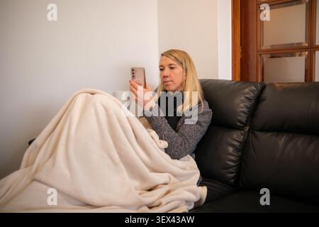 Frau, die auf Sofa mit Decke sitzt, Smartphone benutzt und Freizeit genießt Stockfoto