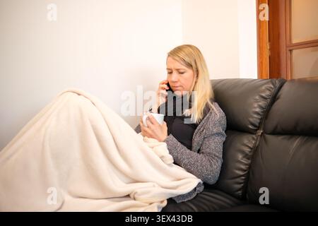 Frau, die sich krank fühlt und auf dem Smartphone einen Becher hält, der auf der Couch sitzt Stockfoto