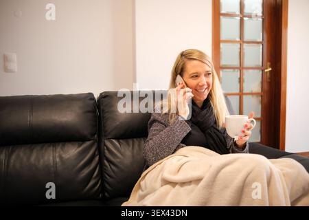 Frau, die sich auf dem Sofa unter der Decke entspannt, Kaffee getrunken und mit dem Handy gesprochen hat Stockfoto