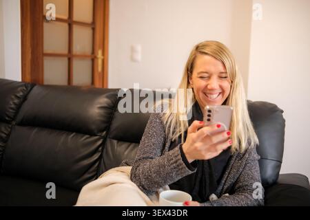 Frau lächelt, während sie sich auf einem Sofa entspannt und auf dem Smartphone nach sozialen Medien und Kommunikation sucht Stockfoto