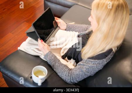 Frau, die sich auf dem Sofa unter der Decke entspannt und die Tablette hält, während sie Tee trinkt Stockfoto
