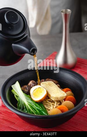 Heiße Brühe in eine Ramen-Schüssel mit Gemüse und Ei gegossen Stockfoto