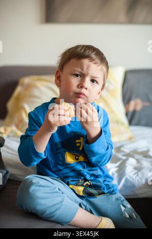 Das kostbare Kind in hellblauer Kleidung schmeckt drinnen eine köstliche Waffel, ein wunderbar ernster Ausdruck, der sein fesselndes Gesicht ziert. Er sitzt komfor Stockfoto