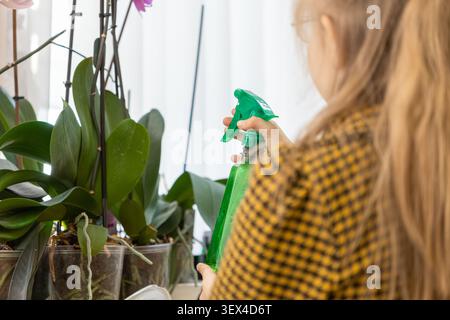 Ein Kind verwendet eine Sprühflasche, um grüne Zimmerpflanzen nahe einem hellen Fenster zu beschlagen, was die Freude an Gartenarbeit, Naturwahrnehmung und Entwicklung von Res widerspiegelt Stockfoto