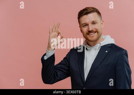 Alles ist in Ordnung. Junger, fröhlicher rothaariger Mann in Freizeitkleidung, der ein gutes Zeichen zeigt und das Leben genießt, glücklich in die Kamera blickt, während er isoliert über rosa Studiohintergrund posiert. Körpersprachkonzept Stockfoto