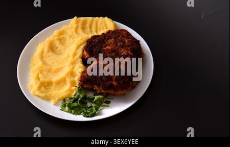 Ein Schweinekotelett, das in Brotkrumen mit Kartoffelpüree auf einem runden Teller vor schwarzem Hintergrund gebraten wird. Draufsicht, flach. Stockfoto