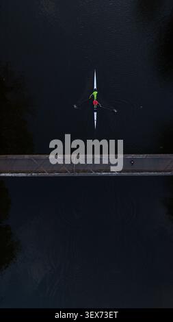 Top-Down-Drohnenaufnahme, die eine minimalistische Szene eines Ruderbootes einfängt, das unter einer Brücke über dunkles Wasser fährt. Stockfoto
