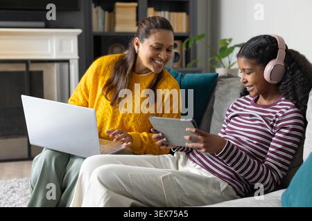 Afroamerikanische Mutter und Jugendtochter sitzen zu Hause auf einem grauen Sofa mit Laptop und Tablet Stockfoto