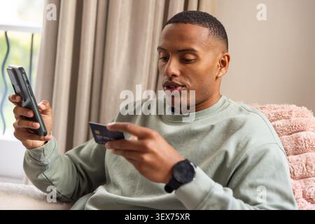 Afrikanischer Mann, der sich zu Hause auf einem hellen Sofa liegend, Smartphone und Karte in der Hand hält und ein grünes Sweatshirt trägt Stockfoto