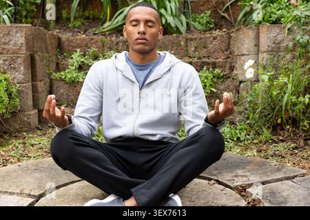 Erwachsener Afroamerikaner sitzend die Beine überkreuzt Meditation im Garten auf Steinplatte mit Hoodie Stockfoto