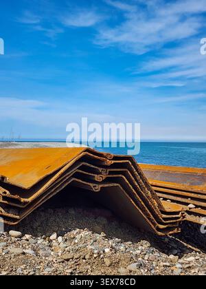 Gestapelte Wellblechpfähle, die auf einem Kiesstrand neben dem Meer liegen, mit Erdbewegungsgeräten und Palmen im Hintergrund, die coa zeigen Stockfoto