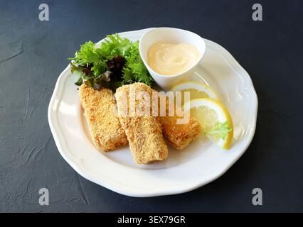 Knusprige panierte Fischfinger serviert mit frischem grünem Salat, Zitronenscheiben und Mayonnaise-Dip auf einem weißen Teller, dunkler Hintergrund. Draufsicht. Stockfoto