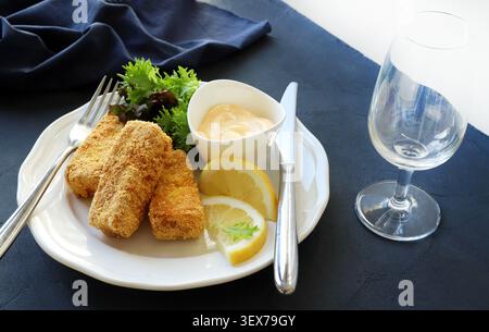 Knusprige panierte Fischfinger serviert mit frischem grünem Salat, Zitronenscheiben, Mayonnaise-Dip auf einem weißen Teller und einem leeren Weinglas. Stockfoto