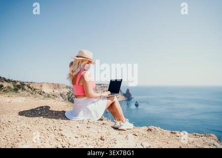 Frau Laptop Freelancer sitzt an einer Klippe und arbeitet ferngesteuert und genießt im Sommer einen wunderschönen Blick aufs Meer Stockfoto
