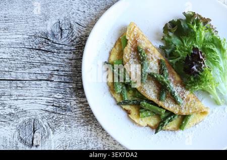 Hausgemachtes Spargel- und Parmesan-Omelette serviert mit frischem grünem Salat auf einem weißen Teller auf einem rustikalen Holzhintergrund. Stockfoto