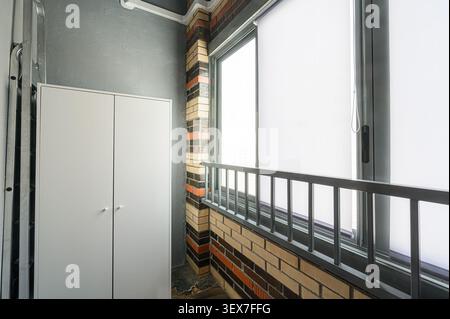 Apartment-Zimmer mit Balkon modernes Interieur mit weißem Schrank an einer grauen Wand, Fenster mit weißen Farbtönen und schwarzem Geländer sowie dekorativen Ziegelsteinen Stockfoto