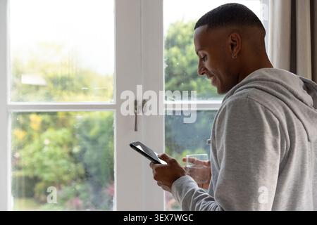 Mann in hellgrauem Hoodie, der an Glastüren steht, Smartphone hält, Glas trinkt, Kopierraum Stockfoto