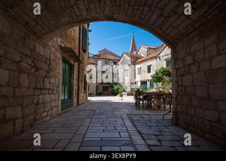 Malerischer Blick auf die Architektur der Altstadt, eingerahmt von einem Steinbogen in Stari Grad, Kroatien. Mediterrane Straße mit Cafétischen, grünen Fensterläden und Bell Stockfoto