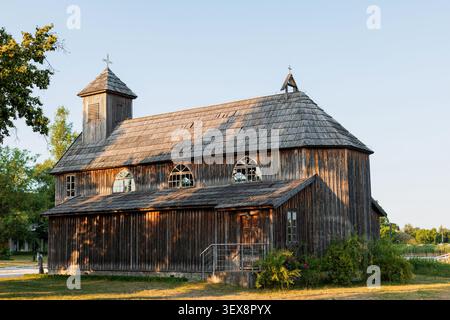 Alte traditionelle Holzkirche mit einem Schindeldach und einem kleinen Turm, beleuchtet von warmem Sonnenuntergangslicht in einer ruhigen ländlichen Gegend unter klarem Himmel Stockfoto