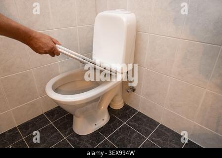 Reinigt die Toilettenschüssel mit einer Bürste von Hand. Weiße Porzellanbefestigung an Fliesenwänden. Dunkle Bodenfliesen stehen im Kontrast zu hellen Wänden. Der Benutzer konzentriert sich auf Sanitat Stockfoto
