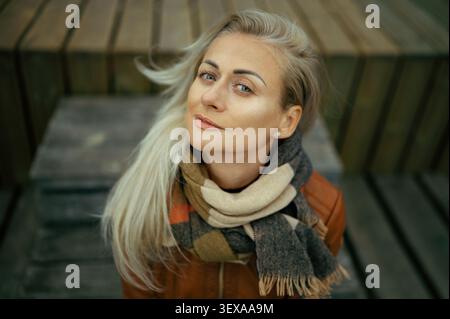Schöne blonde Frau, die lächelt und aus einem hohen Winkel aufblickt, eine Lederjacke und einen Schal auf einem hölzernen Hintergrund trägt Stockfoto