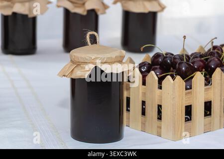 Dunkle hausgemachte Kirschmarmelade in einem Glasgefäß mit Kraftpapierüberzug neben Reifen Kirschen in einer rustikalen Holzkiste, fotografiert als sauberes Stillleben Stockfoto